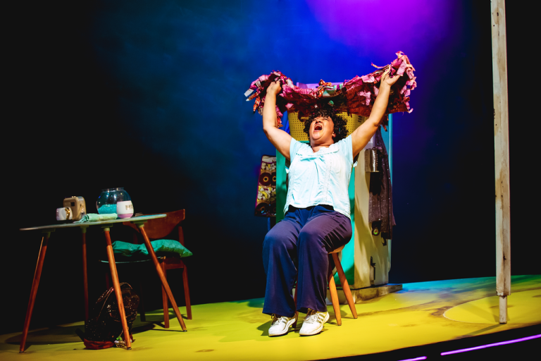 Actor performing on stage with arms above her head with her mouth wide