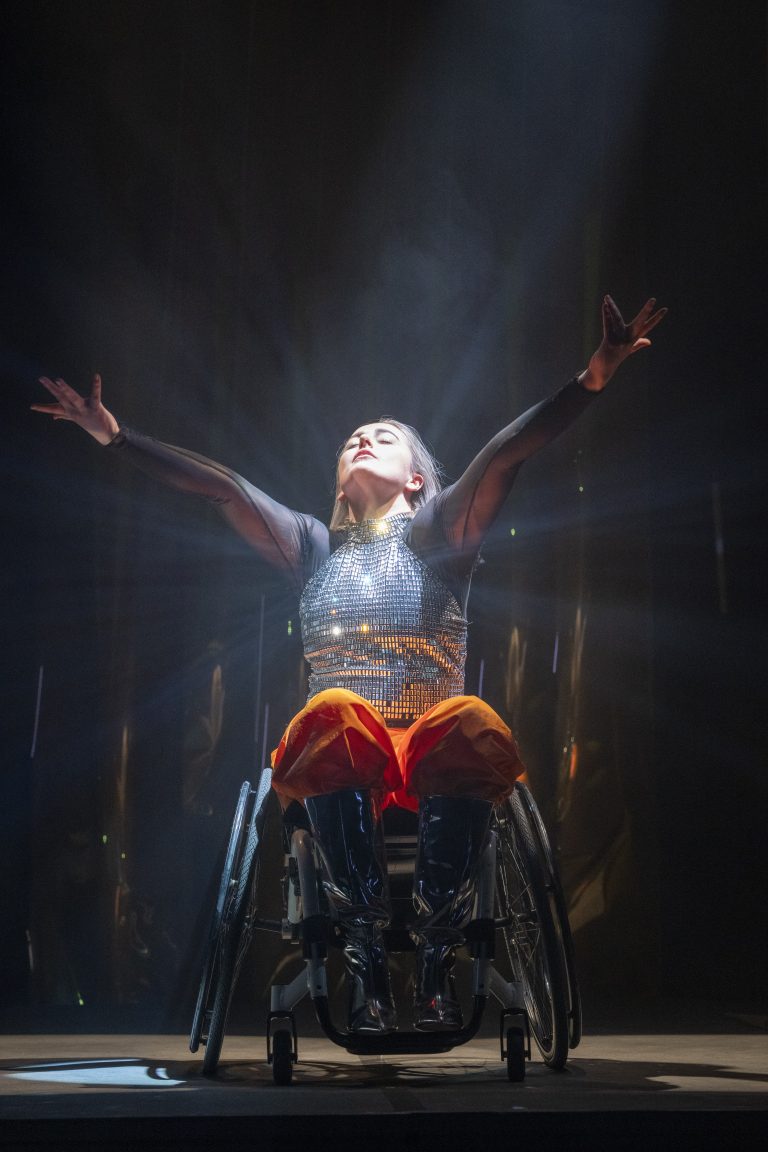 Candoco Dance Company, Over and Over (and over again) by Dan Daw. Photo by Hugo Glendinning. - A woman in a wheelchair dancing with her arms raised under a spotlight, wearing a sparkly silver top and bright orange trousers
